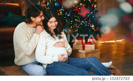 Christmas miracle. Young pregnant woman and her husband sitting by Christmas tree at home together, copy space 132872783