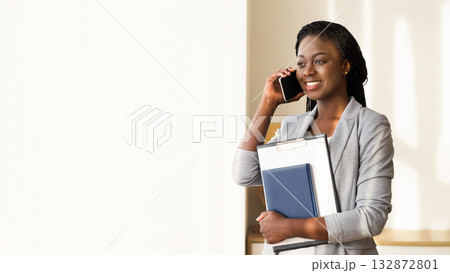 After successful interview. Black female job seeker talking on cellphone and holding folder with CV in her arms, standing near window in office After successful interview. Black female job seeker talking on cellphone and holding folder with CV in her arms, standing near window in office 132872801