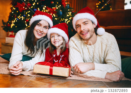 Lovely family of three in red santa hats laying next to Christmas tree, posing on camera, little girl holding xmas gift 132872893