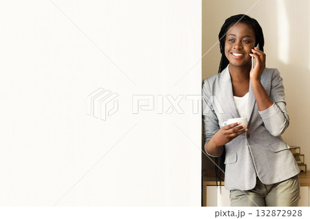 Smiling african american businesswoman talking on cellphone and drinking coffee near window in office, panorama with copy space 132872928