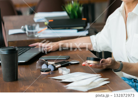 Woman holding cosmetic jar with mineral powderat wooden desk with laptop and coffee cup. Balance between work and self-care, illustrating modern lifestyle and routine. Woman holding cosmetic jar with mineral powderat wooden desk with laptop and coffee cup. Balance between work and self-care, illustrating modern lifestyle and routine. 132872973