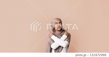 Doubtful black muslim girl in hijab pointing at copy space on yellow background with crossed hands, not sure about her choice 132872990