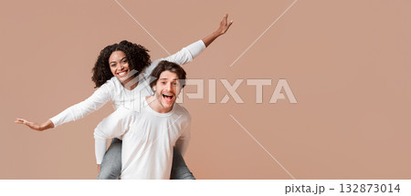 Let's fly. Joyful multiracial couple having fun together over yellow background, afro girl riding her boyfriend's back and spread arms, free space 132873014