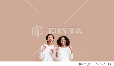 Great offer. Excited multiracial couple looking upwards and pointing at copy space on yellow background above their heads, 132873043