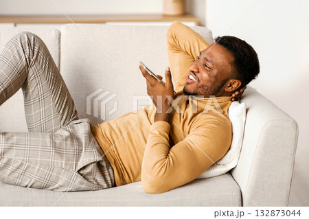 Cheerful African American Guy Using Smartphone Voice Assistant Or Loudspeaker Resting On Couch At Home. 132873044