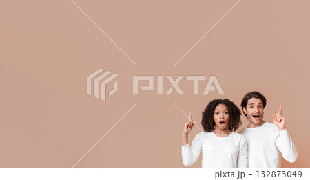 Great Offer. Interracial couple pointing up at copy space above their heads with fingers, posing over yellow background and looking at camera Great Offer. Interracial couple pointing up at copy space above their heads with fingers, posing over yellow background and looking at camera 132873049