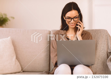 Corporate Communication. Smiling girl talking on mobile phone working on laptop at home, sitting on sofa, copyspace 132873100
