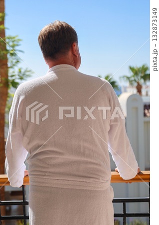 A man in a bathrobe stands on the terrace of a luxury house overlooking the sea and palm trees, rear view 132873149