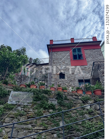 Stone house with red accents and green windows nestled on a hilltop. Decorated with numerous potted plants, it stands against a partly cloudy sky, showcasing rustic architecture. 132874299