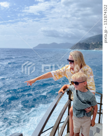 Mother and son standing on a coastal railing enjoy a scenic ocean view together. The mother points outward, both wearing sunglasses and exhibiting happiness and contentment. Mother and son standing on a coastal railing enjoy a scenic ocean view together. The mother points outward, both wearing sunglasses and exhibiting happiness and contentment. 132874322