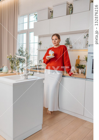 Woman, kitchen, cupcake. Young woman holding a cupcake in a stylish modern white kitchen, enjoying a sweet moment at home. 132876216