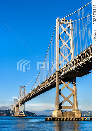 A detailed perspective of the iconic San Francisco-Oakland Bay Bridge's tower and cables, stretching over the bay on a bright day with a clear blue sky. Symbolizes engineering and urban travel A detailed perspective of the iconic San Francisco-Oakland Bay Bridge's tower and cables, stretching over the bay on a bright day with a clear blue sky. Symbolizes engineering and urban travel 132876513