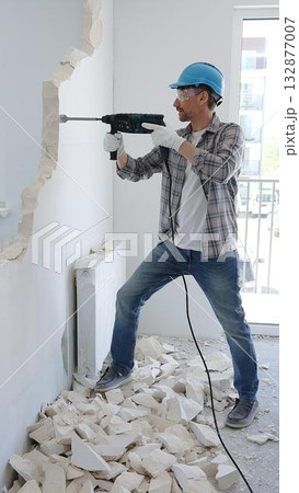 Man construction worker demolishing a wall with a hammer drill in protective gear, creating construction debris on the surrounding floor surface. Renovation concept Man construction worker demolishing a wall with a hammer drill in protective gear, creating construction debris on the surrounding floor surface. Renovation concept 132877007