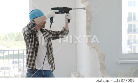Male construction worker demolishing a wall with a hammer drill in protective gear, creating construction debris on the surrounding floor surface. Renovation Male construction worker demolishing a wall with a hammer drill in protective gear, creating construction debris on the surrounding floor surface. Renovation 132877009