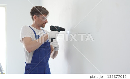 Middle aged male skilled worker drilling hole in white wall, wearing protective gear during home improvement project with cordless power tool 132877013