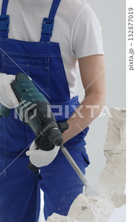 Close up of unknown male construction worker wearing blue overalls is demolishing white wall with rotary hammer drill, generating dust, close up horizontal view. Renovation concept Close up of unknown male construction worker wearing blue overalls is demolishing white wall with rotary hammer drill, generating dust, close up horizontal view. Renovation concept 132877019