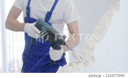 Male construction worker demolishing white wall with rotary hammer drill, wearing blue overalls and protective white gloves, closeup view 132877095