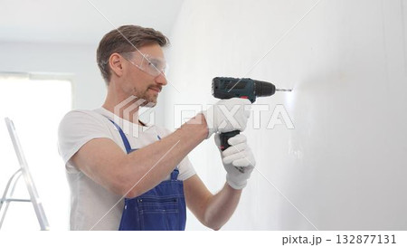Man professional construction worker in a blue jumpsuit, safety goggles, and work gloves is screwing a screw into the wall using a cordless screwdriver, home renovation 132877131
