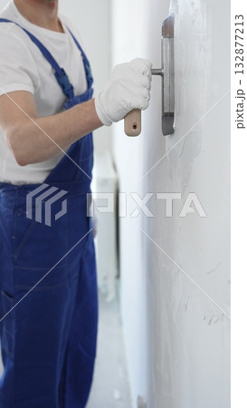 Professional construction worker wearing blue work overall is using a finishing trowel to carefully apply plaster on a wall, demonstrating expertise in home renovation and interior finishing 132877213