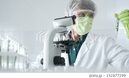 Adult male scientist wearing green protective gear looking at plant samples in test tubes and using a microscope in modern laboratory for botanical research. Medicine, healthcare and science concept Adult male scientist wearing green protective gear looking at plant samples in test tubes and using a microscope in modern laboratory for botanical research. Medicine, healthcare and science concept 132877299