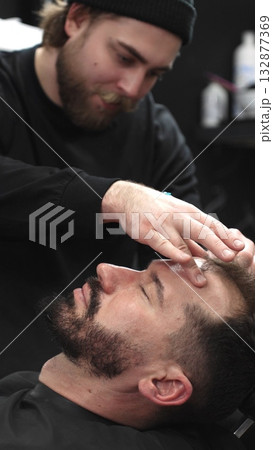 Professional barber carefully shaving client's beard with straight razor, applying gel and using hot towel for precise grooming in contemporary barbershop setting, vertical view 132877369