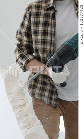Close up of unknown male construction worker wearing beige checkered shirt and protective gloves, is demolishing white wall with rotary hammer drill, generating dust. Renovation project Close up of unknown male construction worker wearing beige checkered shirt and protective gloves, is demolishing white wall with rotary hammer drill, generating dust. Renovation project 132877428