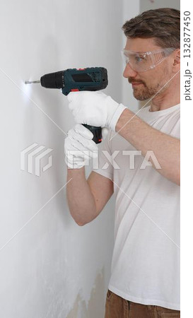Middle aged male worker wearing white t-shirt and protective gloves, is drilling hole in a wall with cordless drill during home renovation work. Portrait view 132877450