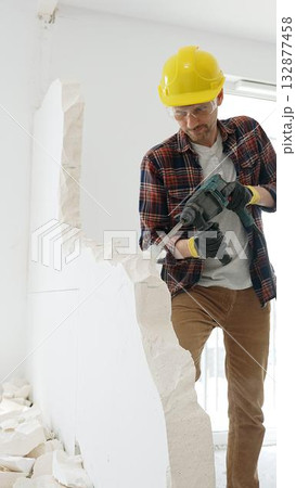 Male construction worker wearing red checkered shirt, yellow protective helmet, is demolishing white wall with rotary hammer drill, generating dust, medium long shot. Renovation project Male construction worker wearing red checkered shirt, yellow protective helmet, is demolishing white wall with rotary hammer drill, generating dust, medium long shot. Renovation project 132877458