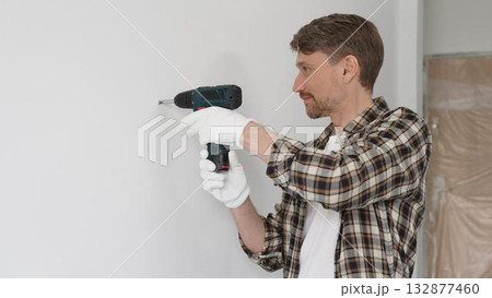 Man construction worker in checked shirt and safety gloves is screwing a screw into the wall using a cordless screwdriver, carrying out a careful home renovation. Horizontal portrait view Man construction worker in checked shirt and safety gloves is screwing a screw into the wall using a cordless screwdriver, carrying out a careful home renovation. Horizontal portrait view 132877460