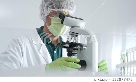 Medical scientist wearing protective mask, cap and green gloves using microscope doing scientific research in laboratory, developing vaccine or medication. Medicine, healthcare and science concept 132877473