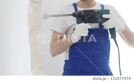 Close up of unrecognizable male construction worker wearing blue overalls and protective white gloves, is demolishing white wall with rotary hammer drill, generating dust, horizontal view. Renovation 132877474