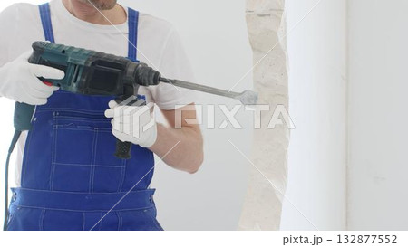 Unrecognizable male construction worker wearing blue overalls and protective white gloves, is demolishing white wall with rotary hammer drill, generating dust, close up horizontal view. Renovation 132877552
