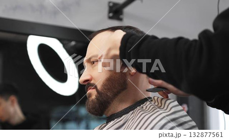 Skilled barber carefully grooming client's beard with electric razor, creating precise, trendy look inside contemporary barbershop setting 132877561