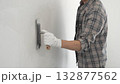 Unknown male construction worker wearing gloves and plaid shirt is using finishing trowel to plaster a white wall, applying a layer of plaster to create a smooth surface during renovation project 132877562