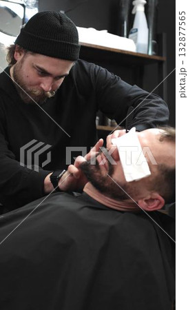 Professional barber carefully shaving client's beard with straight razor, applying gel and using hot towel in barbershop setting, vertical view Professional barber carefully shaving client's beard with straight razor, applying gel and using hot towel in barbershop setting, vertical view 132877565