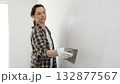 Woman construction professional wearing checkered shirt is smoothing white wall surface, applying putty with professional spatula technique, showcasing skilled interior renovation work 132877567