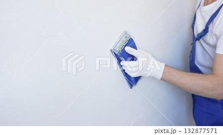 Unknown male construction worker wearing protective white gloves and blue construction coveralls, is sanding white wall with blue hand block, preparing smooth surface during interior home renovation Unknown male construction worker wearing protective white gloves and blue construction coveralls, is sanding white wall with blue hand block, preparing smooth surface during interior home renovation 132877571
