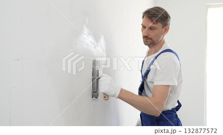 Professional male construction worker is using a finishing trowel to carefully apply plaster on a wall, demonstrating expertise in home renovation and interior finishing. Portrait view Professional male construction worker is using a finishing trowel to carefully apply plaster on a wall, demonstrating expertise in home renovation and interior finishing. Portrait view 132877572