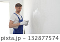 Middle aged male construction worker wearing gloves and blue overalls is using a putty knife to apply plaster on a white wall, performing home renovation 132877574