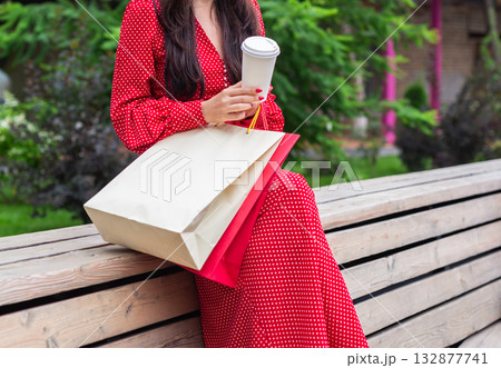 Woman in red polka dot dress sitting with coffee cup and shopping bags outdoors. Relaxation, consumer lifestyle, and leisure concept. 132877741