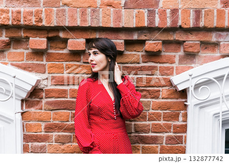 Woman in red dress standing near vintage brick wall and smiling. Confidence, beauty, and timeless urban style. 132877742