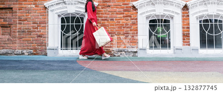 Woman in red polka dot dress walking with shopping bag near vintage brick building Banner with copy space and empty place for advertising. Fashion, city lifestyle, and elegance in urban environment. Woman in red polka dot dress walking with shopping bag near vintage brick building Banner with copy space and empty place for advertising. Fashion, city lifestyle, and elegance in urban environment. 132877745