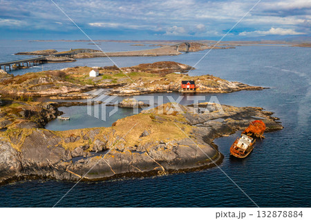 Rusty shipwreck partially submerged in calm coastal waters near a red boathouse on a rocky islet. The Atlantic Ocean Road stretches across Norways wild and rugged seascape Rusty shipwreck partially submerged in calm coastal waters near a red boathouse on a rocky islet. The Atlantic Ocean Road stretches across Norways wild and rugged seascape 132878884