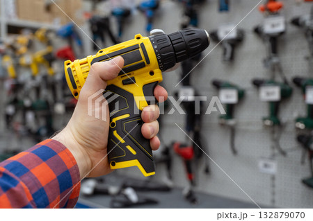 A man in a hardware store chooses a new drill or screwdriver. A hand holds a yellow power drill in front of a hardware store display with various tools. Handyman holding electric screwdriver. 132879070