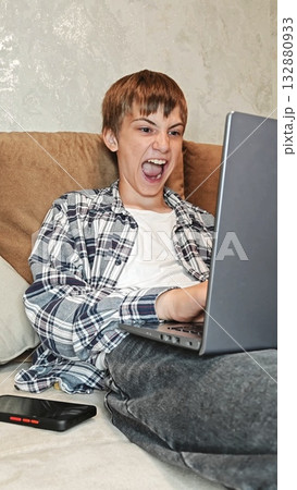 Young man at home smiling with satisfaction, is emotionally working on a laptop. Caucasian guy sitting on sofa and enthusiastically taps on the keyboard. crazy 132880933