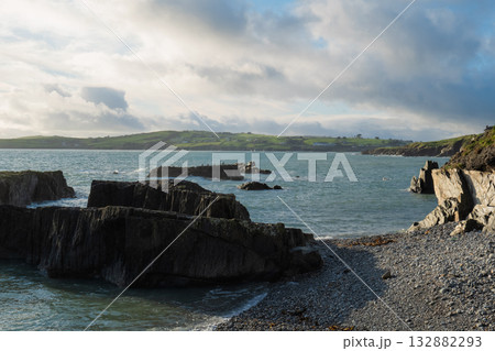 Gentle waves lap against dark rocks and a pebble beach at Sheep Cove. Green hills and a cloudy sky fill the background on a bright day in Ireland. 132882293