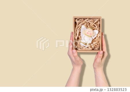 Hands Presenting Christmas Ornament Gift Box With Angel Figurine On Beige Background. Concept Of Holiday Decorations, Festive Packaging Ideas, Christmas Gifting Trends, And Seasonal Home Styling Hands Presenting Christmas Ornament Gift Box With Angel Figurine On Beige Background. Concept Of Holiday Decorations, Festive Packaging Ideas, Christmas Gifting Trends, And Seasonal Home Styling 132883523