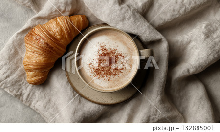 Minimalist flat lay of cappuccino with cinnamon and croissant on linen napkin, neutral coffeehouse aesthetic for lifestyle content. 132885001
