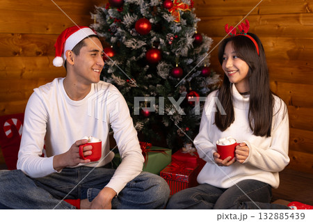 A young couple sits together in front of a Christmas tree, smiling A young couple sits together in front of a Christmas tree, smiling 132885439