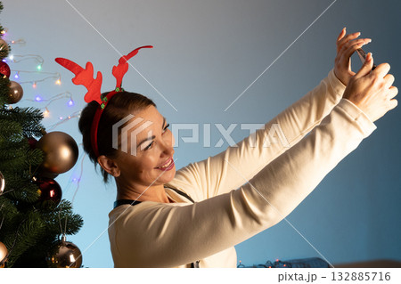woman wearing reindeer antlers is taking a selfie next to a decorated Christmas woman wearing reindeer antlers is taking a selfie next to a decorated Christmas 132885716